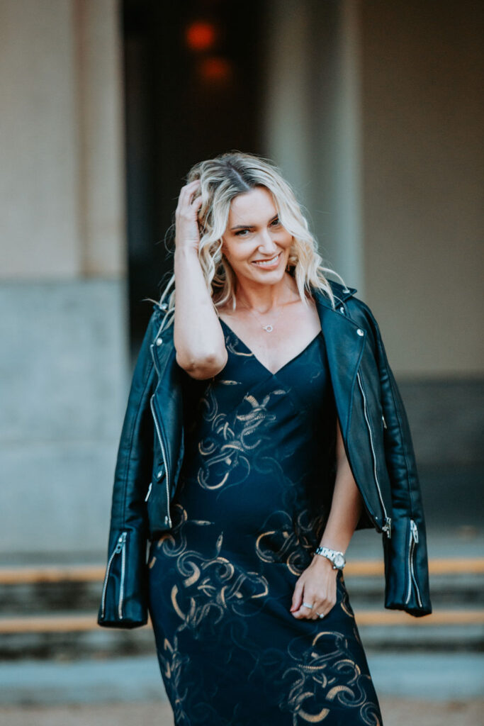 Personal stylist Jaquelyn Wahidi wears a black and gold slip dress and leather jacket in front of a civic building.