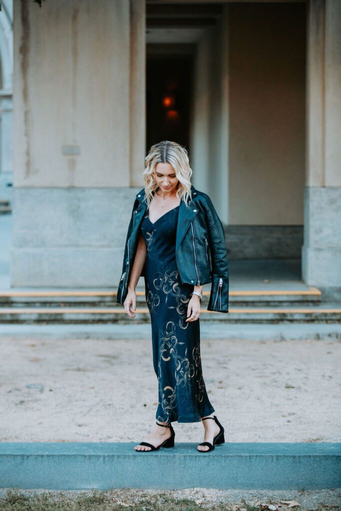 Personal stylist Jaquelyn Wahidi wears a black and gold slip dress with a leather jacket in front of an art center.